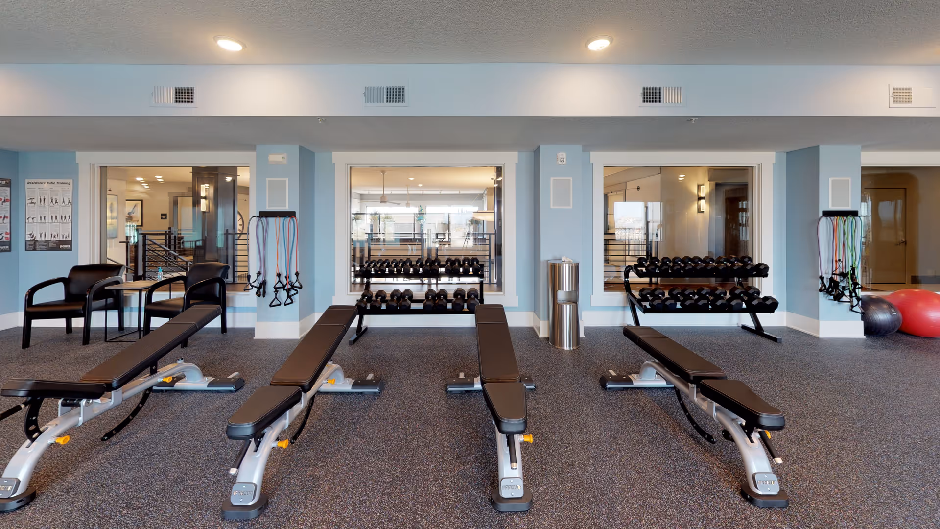 Interior view of a fitness room with four black adjustable weight benches arranged in a row. Behind the benches are racks filled with dumbbells and resistance bands hanging on the walls. Two black chairs and a small table are positioned on the left side. The room has light blue walls, large windows, and overhead lighting.