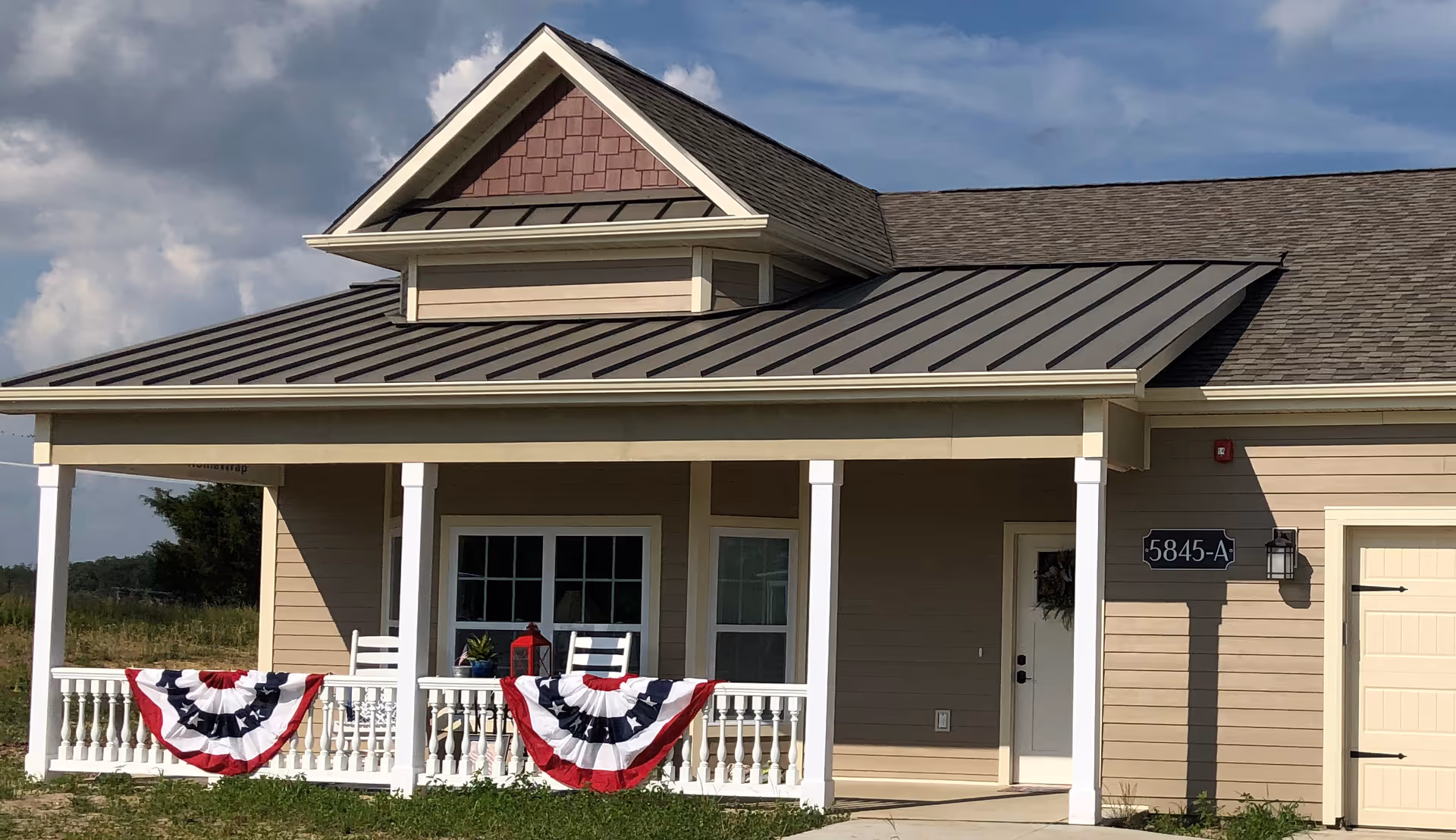 Exterior view of a single-story beige building with a covered porch decorated with red, white, and blue bunting. The porch has white railings and two white rocking chairs. The building has a gray metal roof and a sign near the door that reads '5845-A'.