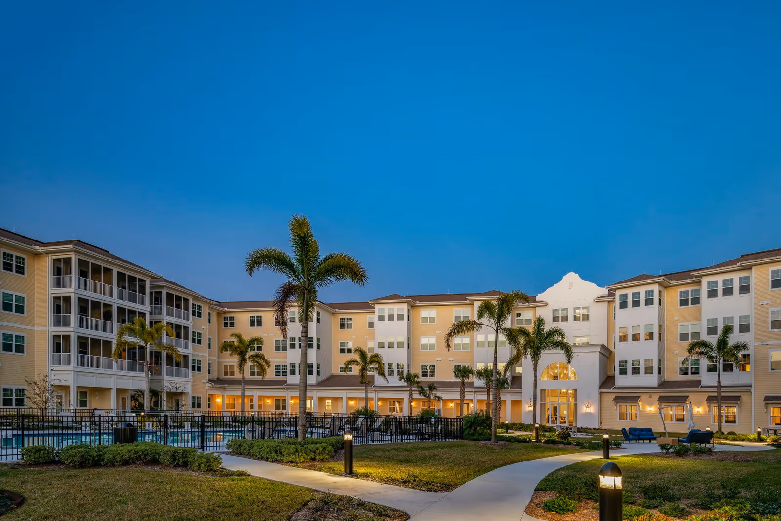 Exterior view of Arbor Terrace Citrus Park Independent Living facility at dusk, showing a large multi-story building with balconies, palm trees, a swimming pool area enclosed by a fence, and well-lit pathways surrounded by landscaped greenery.