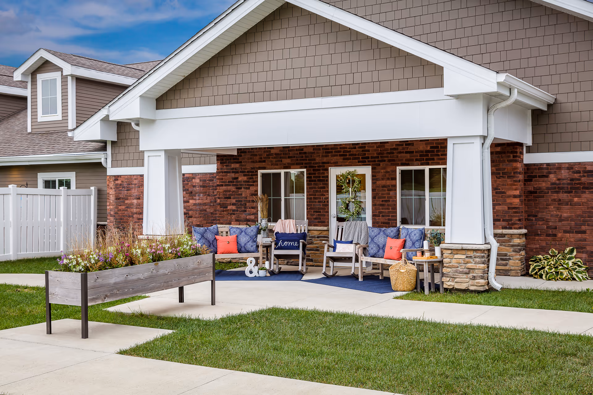 Outdoor seating area at a senior living facility with wooden rocking chairs and a bench with blue and red cushions, a small table with decorative items, and a planter box with flowers. The area is covered by a roof attached to a building with brick and siding exterior walls.