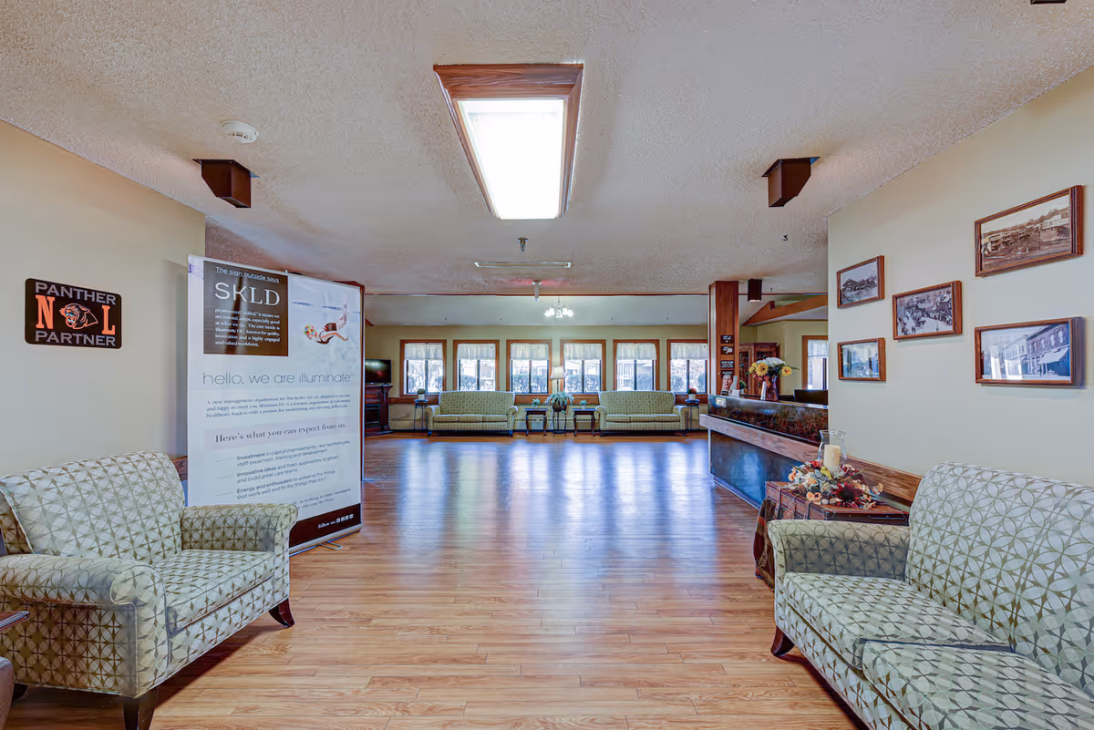 A spacious living room area in a senior living facility with wooden flooring, patterned armchairs on both sides, and a row of green sofas near large windows at the far end. The walls are decorated with framed pictures and a sign that reads 'Panther N L Partner.' A large informational banner stands near the left side, and a wooden counter with floral decorations is on the right.