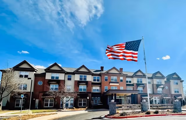 Exterior view of a three-story senior living facility building with a large American flag flying on a flagpole in front. The building has a combination of brick and light-colored siding with multiple windows and gabled roofs. The sky is clear with a few clouds.