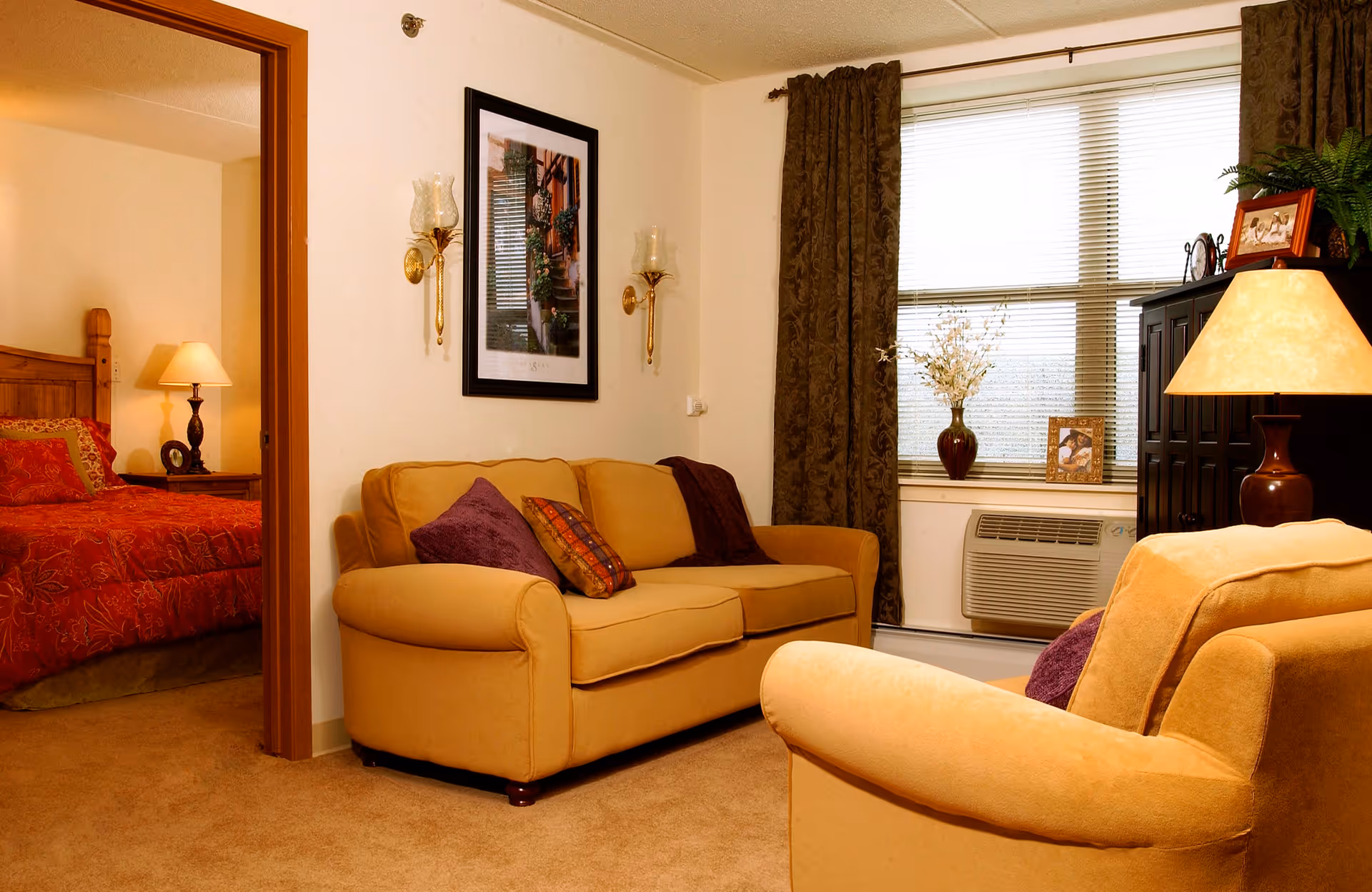 A cozy living room with a beige sofa and armchair, decorated with purple and patterned throw pillows. There are two wall sconces and a framed picture on the wall. A window with brown curtains and blinds lets in natural light. A dark wooden cabinet with a lamp, framed photos, and a plant is next to the window. Through an open doorway, a bedroom with a wooden bed frame, red bedding, and a bedside lamp is visible.
