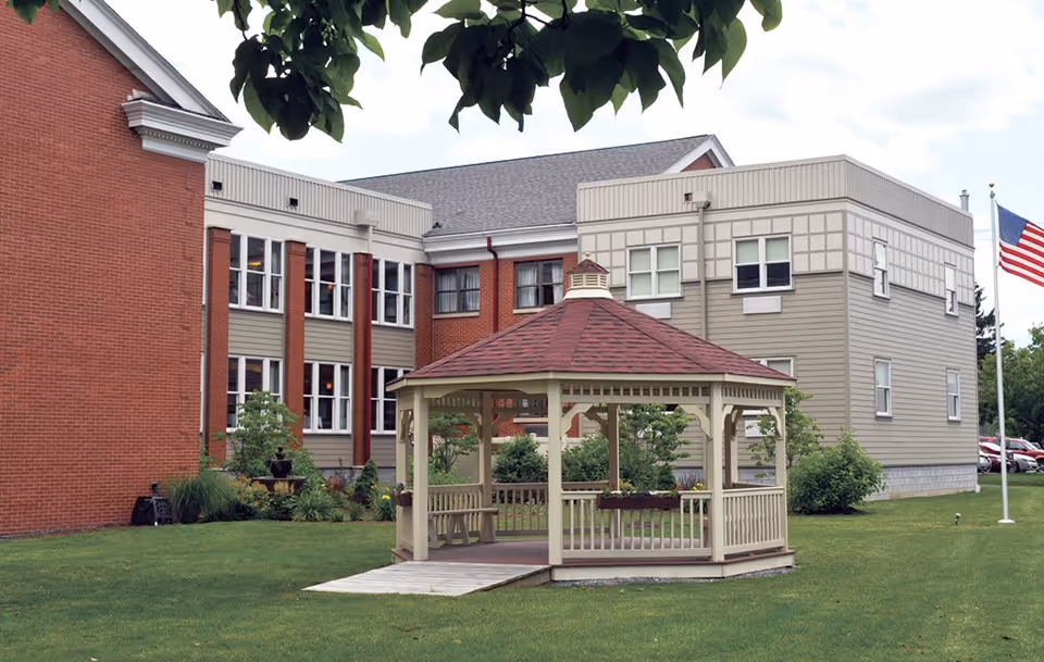 Outdoor view of Presbyterian Village at Hollidaysburg showing a beige gazebo with a red roof on a grassy lawn. The building in the background has red brick and beige siding with multiple windows. An American flag is visible on a flagpole to the right.
