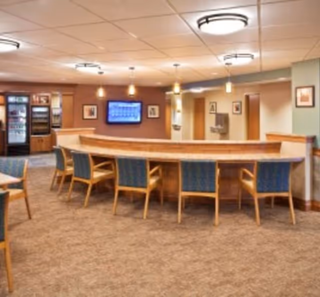 Communal dining area with a long curved counter lined with chairs, a wall-mounted TV, and a refrigerated display in the background.