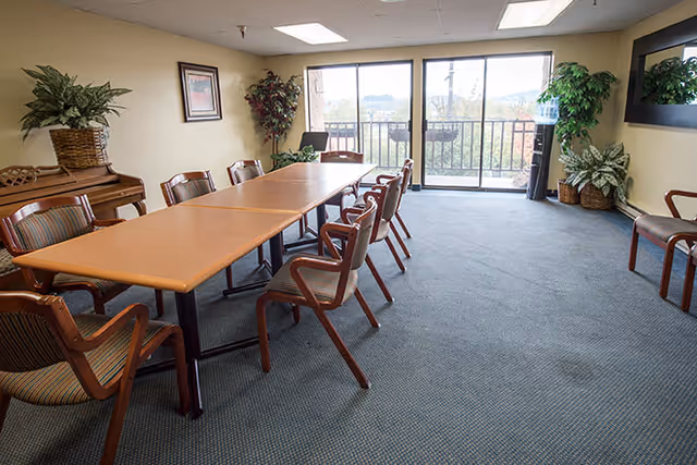 A meeting or dining room with a long rectangular table surrounded by wooden chairs with striped cushions. The room has large windows and a glass door leading to a balcony with a view outside. There are several potted plants and a piano against one wall.