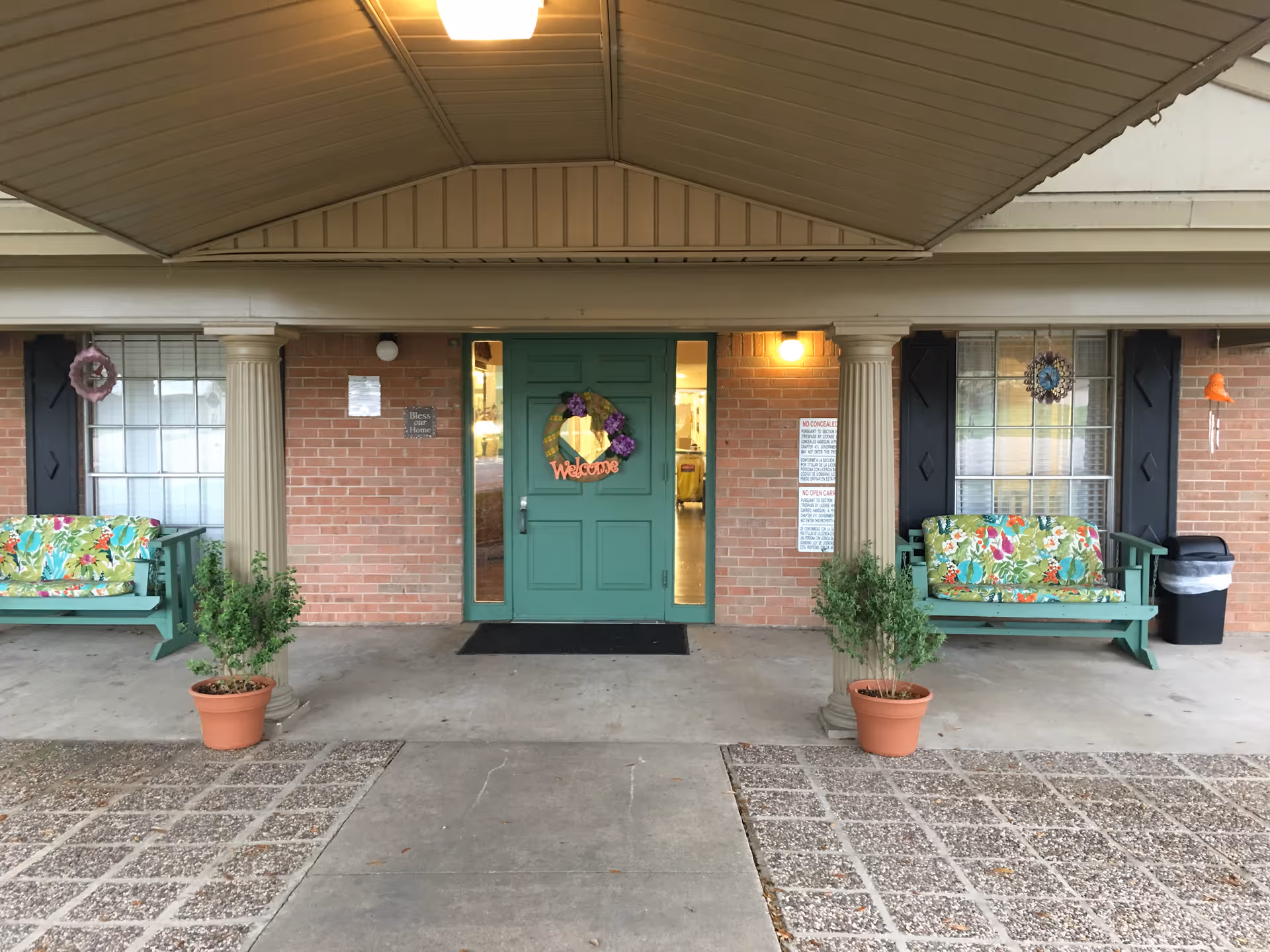 Covered front entrance of a brick assisted living building with a green double door decorated with a wreath, floral-cushioned benches, and potted plants.