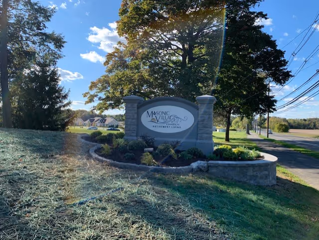 Outdoor view of the entrance sign for Masonic Village Retirement Living, surrounded by landscaping with trees, grass, and a clear blue sky with some clouds.