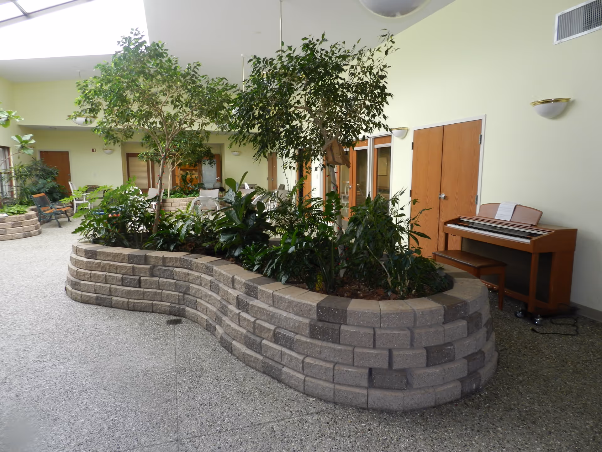 Indoor common area with a large curved planter filled with various green plants and small trees. The space has light yellow walls, a piano with a bench on the right side, and several doors and windows along the walls. There are also chairs and benches placed around the area, and the floor is covered with speckled terrazzo.