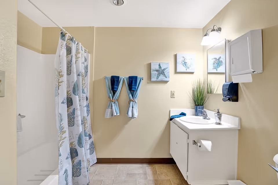 A clean bathroom with beige walls featuring a white bathtub with a shower curtain decorated with seashells and marine life. Two blue and white towels are neatly hung on the wall. There is a white sink vanity with a mirror above it, a soap dispenser, a paper towel dispenser, and a potted plant. Two small marine-themed paintings hang on the wall above the towels.