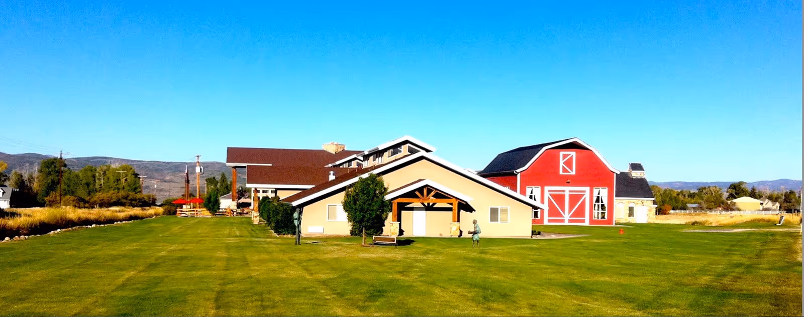 Wide exterior view of a senior living property with a large green lawn, a low beige building with a wooden entrance, and a red barn under a clear blue sky.