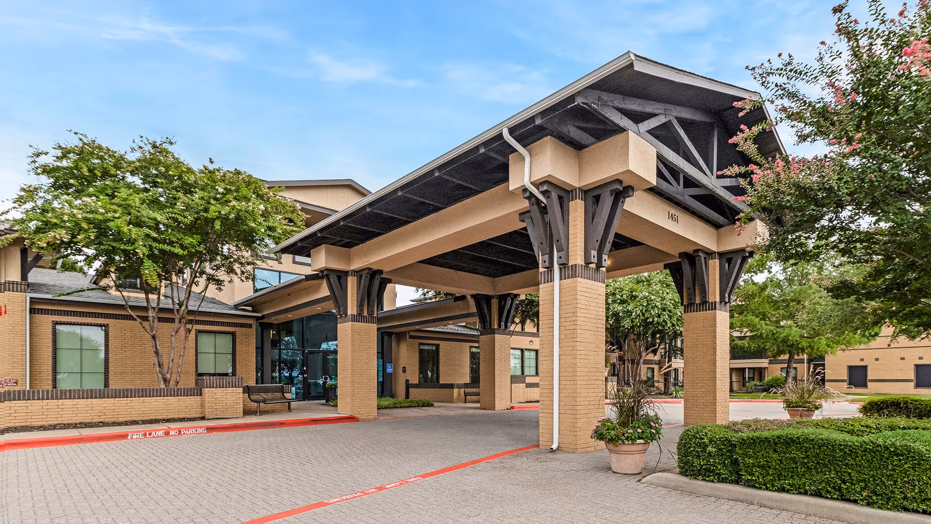 Entrance of a senior living facility with a covered drop-off area supported by large pillars. The building is made of light brown brick with large windows and surrounded by trees and shrubs under a partly cloudy sky.