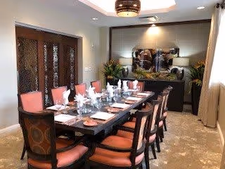 A formal dining room with a long dark wooden table set for twelve people. The table is arranged with white napkins, plates, glasses, and silverware. The chairs have dark wood frames with coral-colored cushions. The room features a decorative light fixture on the ceiling, a sideboard with plants and artwork on the wall behind it, and large windows with beige curtains allowing natural light in.
