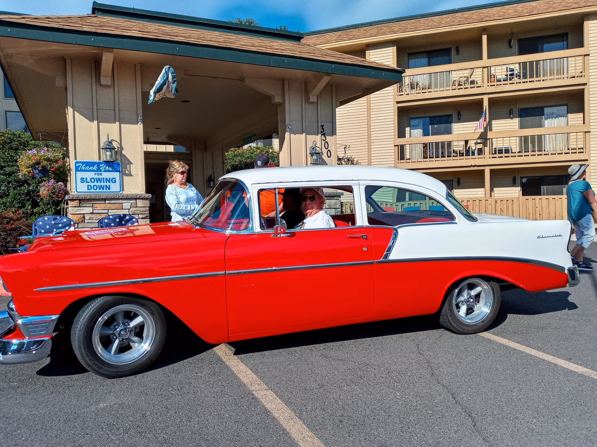 A vintage red and white Chevrolet car parked in front of a building entrance with people around it. The building has balconies and an American flag is visible. A sign near the entrance reads 'Thank You for Slowing Down.'