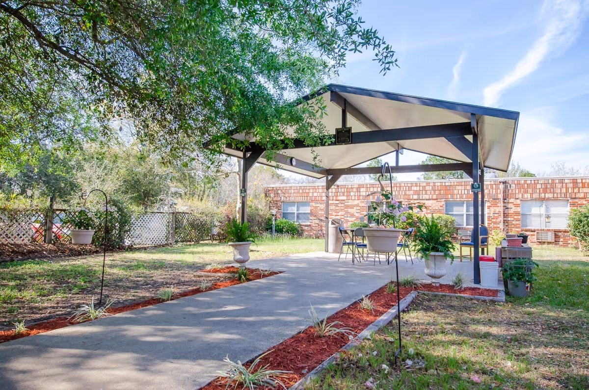 Covered outdoor pavilion with a concrete walkway, patio chairs, tables, and potted plants in a grassy courtyard in front of a brick building.