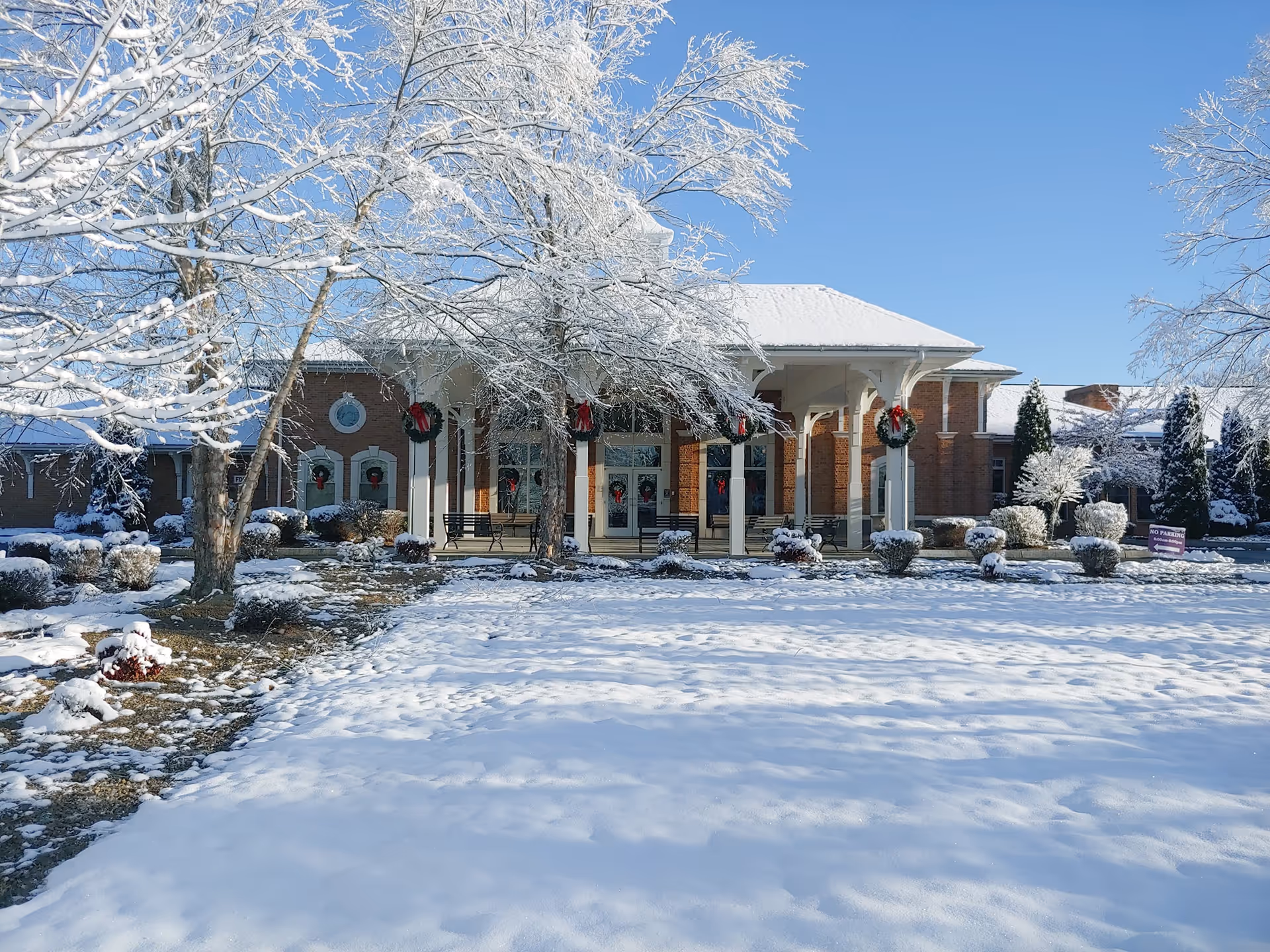 Snow-covered lawn and trees in front of a brick retirement facility entrance decorated with wreaths.
