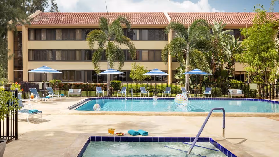 Outdoor swimming pool area at American House Sarasota with several blue and white striped umbrellas, lounge chairs, palm trees, and a hot tub in the foreground. The background shows a three-story building with many windows.