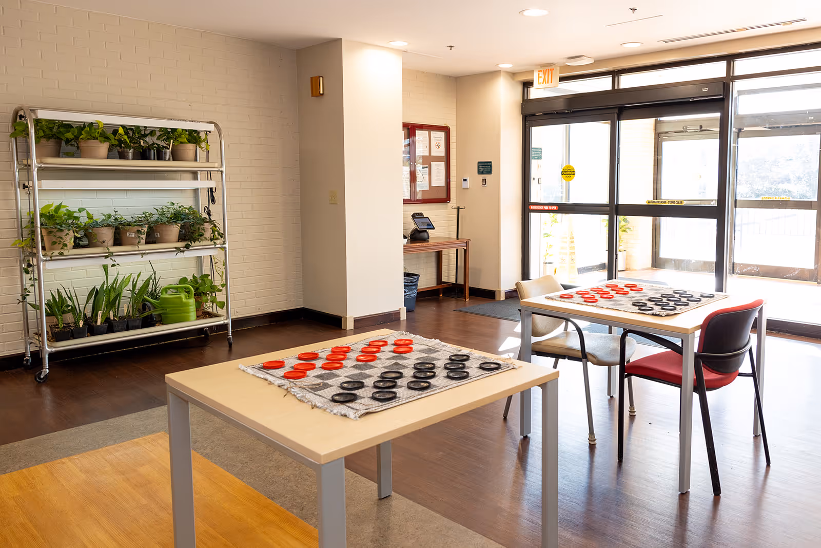 A bright room with two tables set up for checkers games, each with red and black pieces on a checkered cloth. There are two chairs at one table and one chair at the other. A metal rack with several potted plants and a green watering can is against a white brick wall. The room has large glass doors letting in natural light and an exit sign above the door.