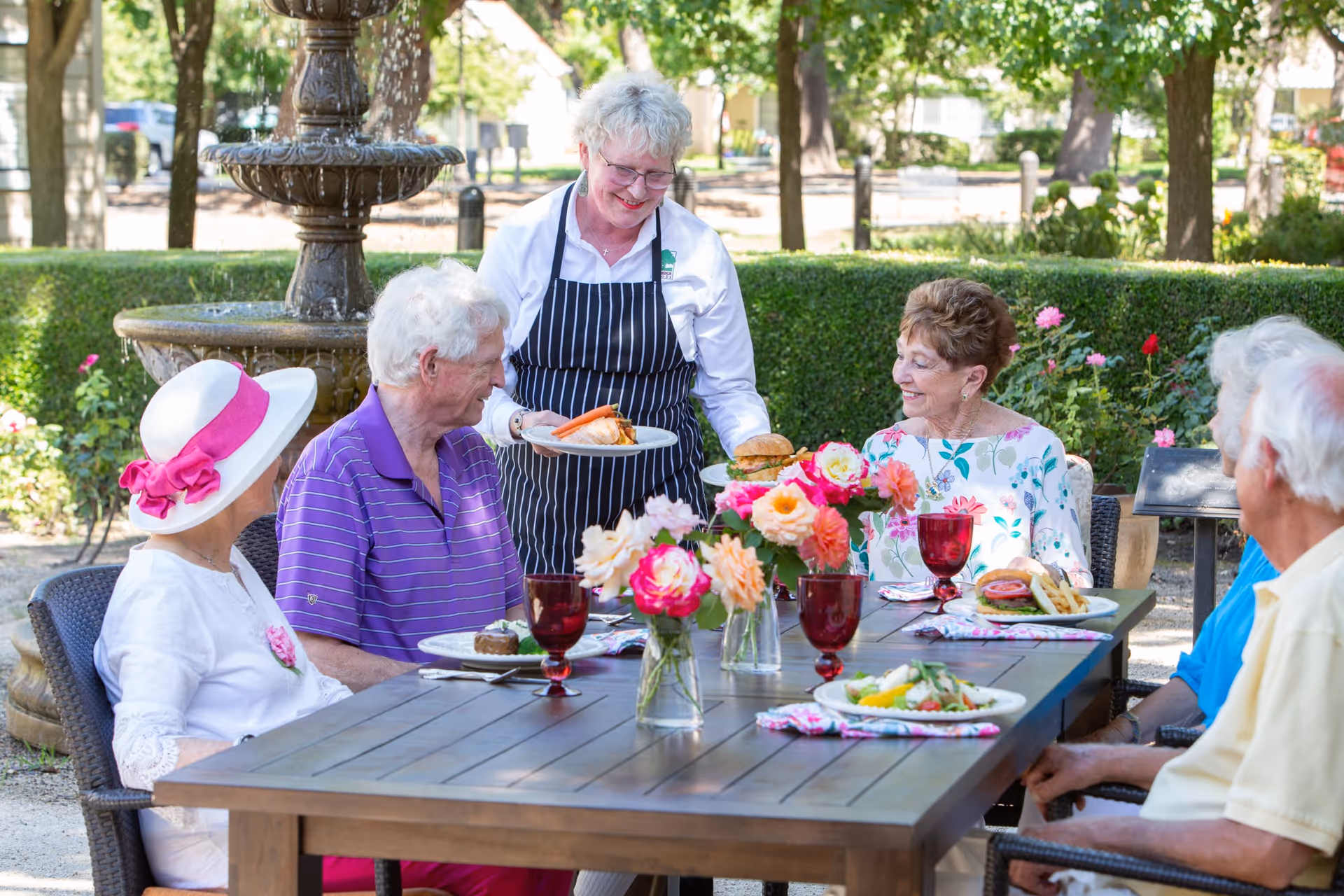 A group of elderly people sitting around a wooden outdoor dining table with flowers in vases, being served food by a smiling woman in a striped apron. The setting is a garden with a fountain and greenery in the background.