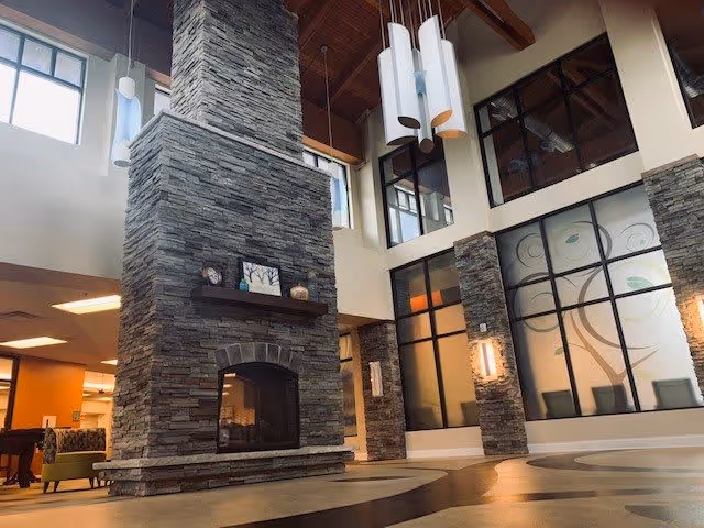 Interior view of a spacious senior living facility common area with a tall stone fireplace in the center, large windows, modern pendant lights hanging from a high wooden ceiling, and decorative wall art featuring a tree design.