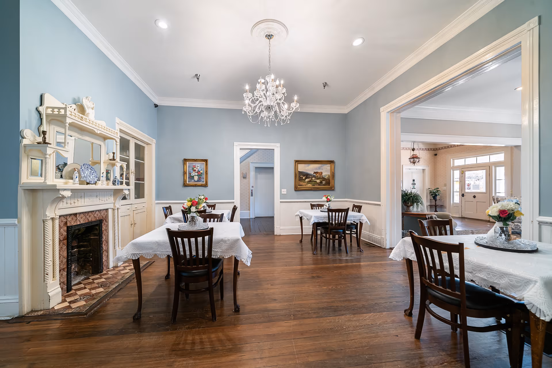 A dining room with wooden floors and blue walls featuring a white ornate fireplace on the left. There are several tables covered with white lace tablecloths, each with a small floral centerpiece. A crystal chandelier hangs from the ceiling. The room opens into another area with a door and windows visible in the background.