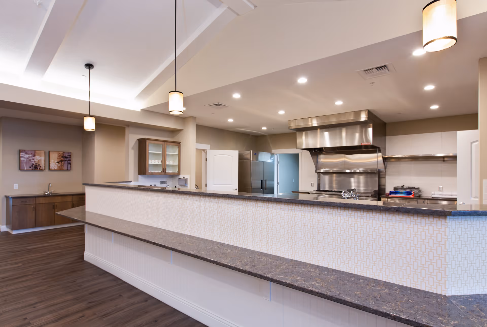 A modern commercial kitchen with stainless steel appliances including a large stove and refrigerator. The kitchen is separated from the dining or serving area by a long counter with a patterned front and dark countertop. The room features wood flooring, pendant lights hanging from the ceiling, and a small sink area with cabinets and wall art in the background.