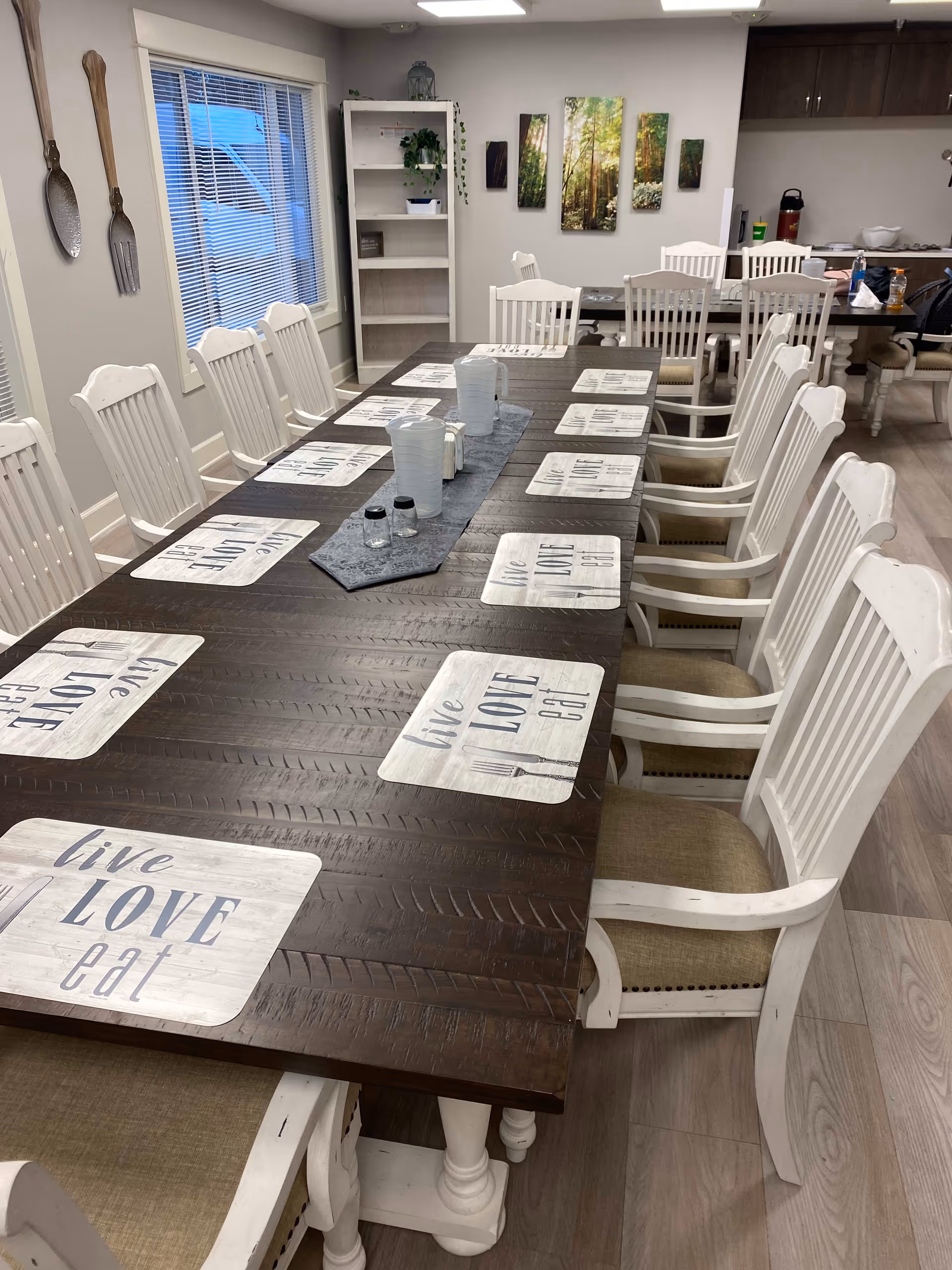 A long dark wooden dining table set with white placemats that say 'live LOVE eat' and white pitchers and salt and pepper shakers in the center. The table is surrounded by white wooden chairs with beige cushions. The room has light-colored walls, a window with blinds, wall decorations including large wooden spoon and fork, and a multi-panel forest-themed artwork. There is a white bookshelf and a kitchen counter with cabinets in the background.