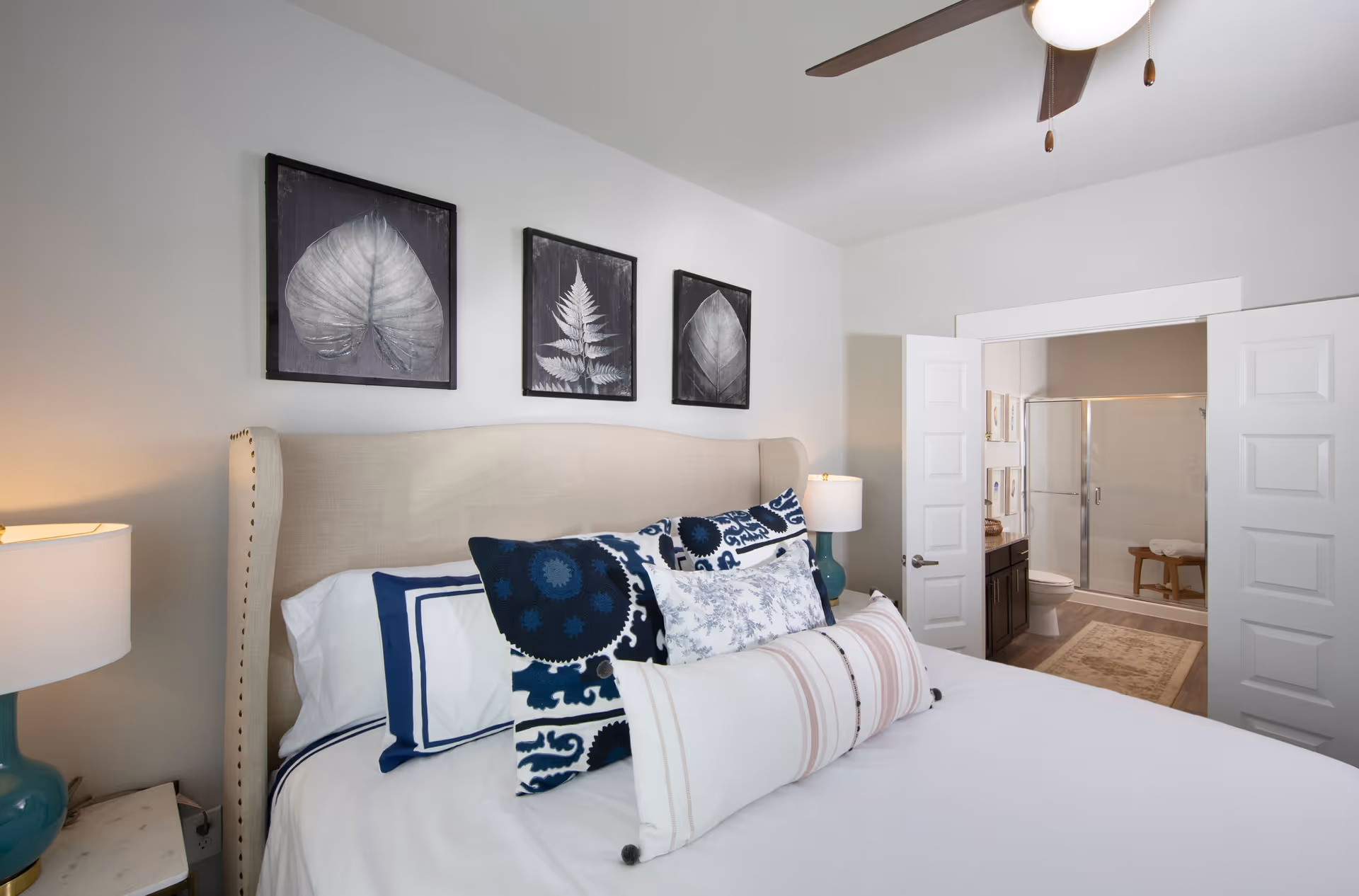 A bedroom with a beige upholstered headboard and a neatly made bed with multiple decorative pillows in blue, white, and beige. Above the bed are three framed black and white leaf prints. On either side of the bed are nightstands with teal lamps. In the background, double doors open to reveal a bathroom with a glass shower door, a toilet, and a vanity with a dark cabinet and a rug on the floor.