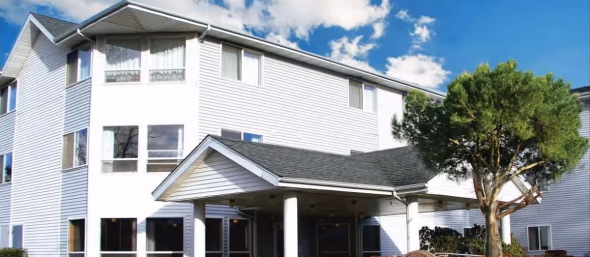 Exterior view of a multi-story senior living facility building with white siding, multiple windows, a covered entrance supported by white columns, and a tree in the foreground under a partly cloudy blue sky.