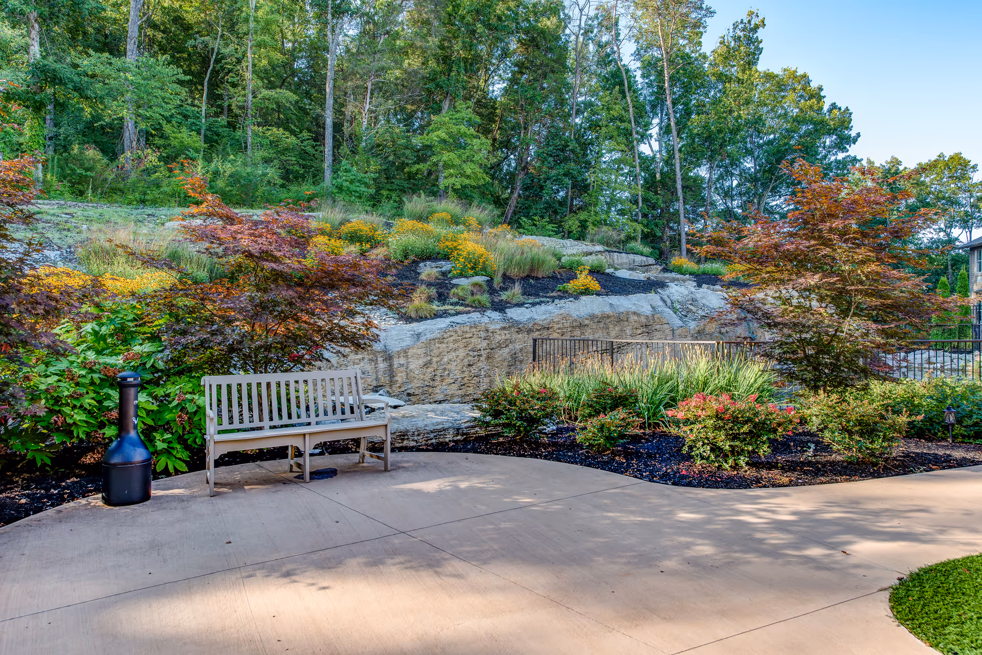 Outdoor garden area with a wooden bench and a black chiminea on a paved patio. Surrounding the patio are various shrubs, flowering plants, and small trees with a rocky hillside and tall trees in the background under a clear blue sky.