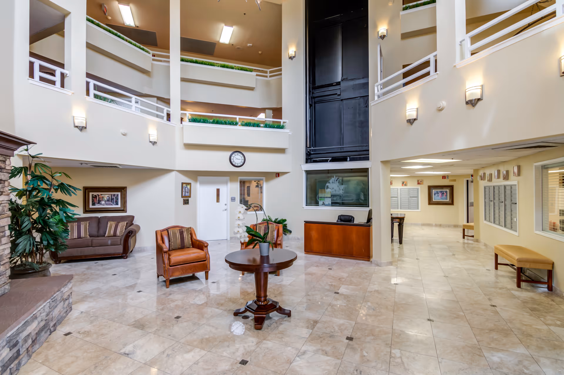 A spacious and well-lit interior common area of a senior living facility with a high ceiling and multiple levels visible. The room features a stone fireplace on the left, a brown sofa, a leather armchair, a round wooden table with a potted plant, and a reception desk. The floor is tiled with a polished marble pattern, and there are several wall-mounted lights and framed pictures. A clock is mounted on the wall above a door, and there are railings on the upper levels overlooking the area.