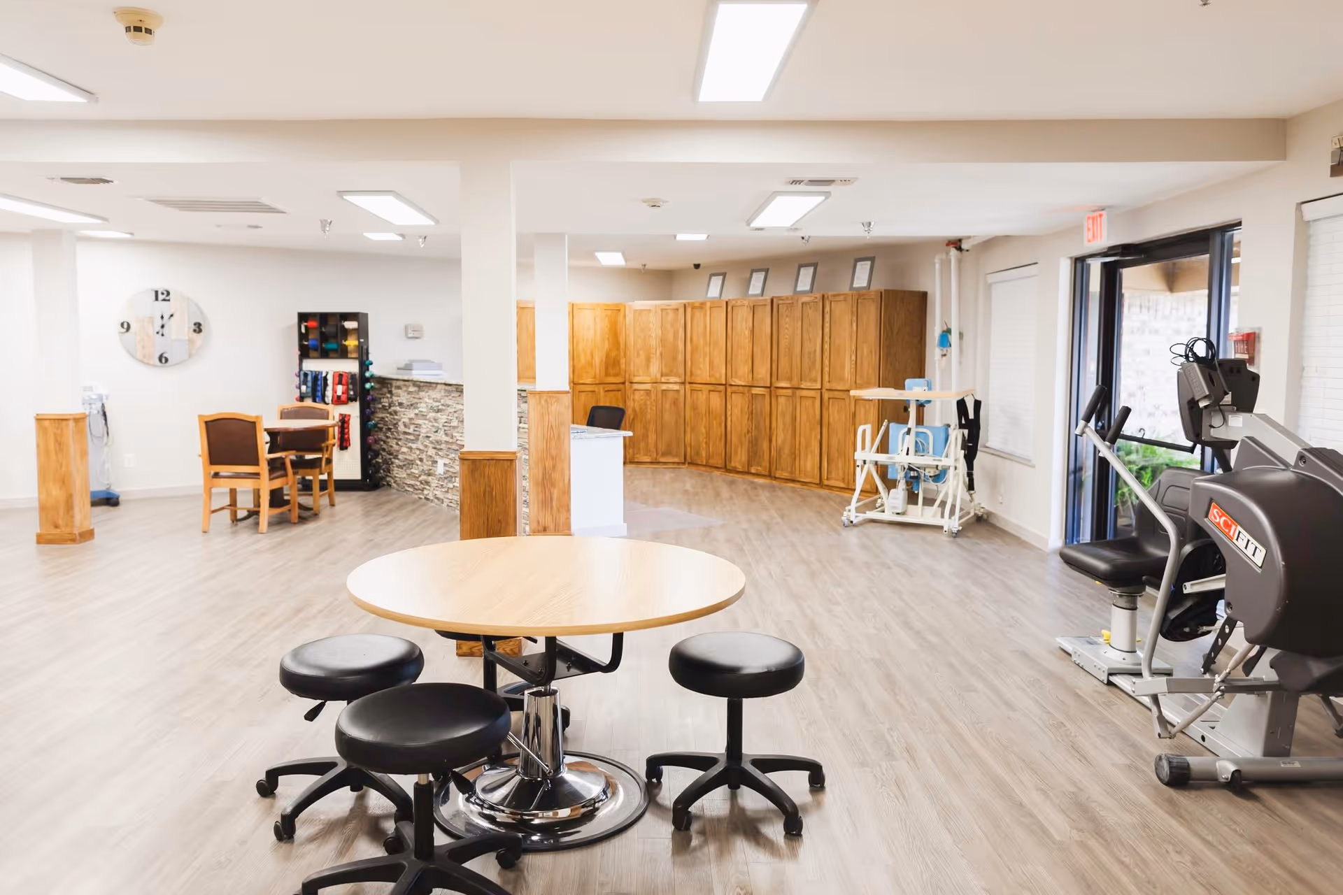 Bright communal activity and therapy room with a round table and stools, exercise equipment, wooden cabinets, and seating.