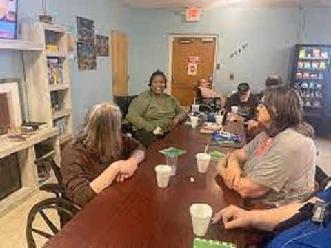 A group of people sitting around a long wooden table in a room with light blue walls. There are cups and some papers on the table. Shelves with books and a vending machine are visible in the background.