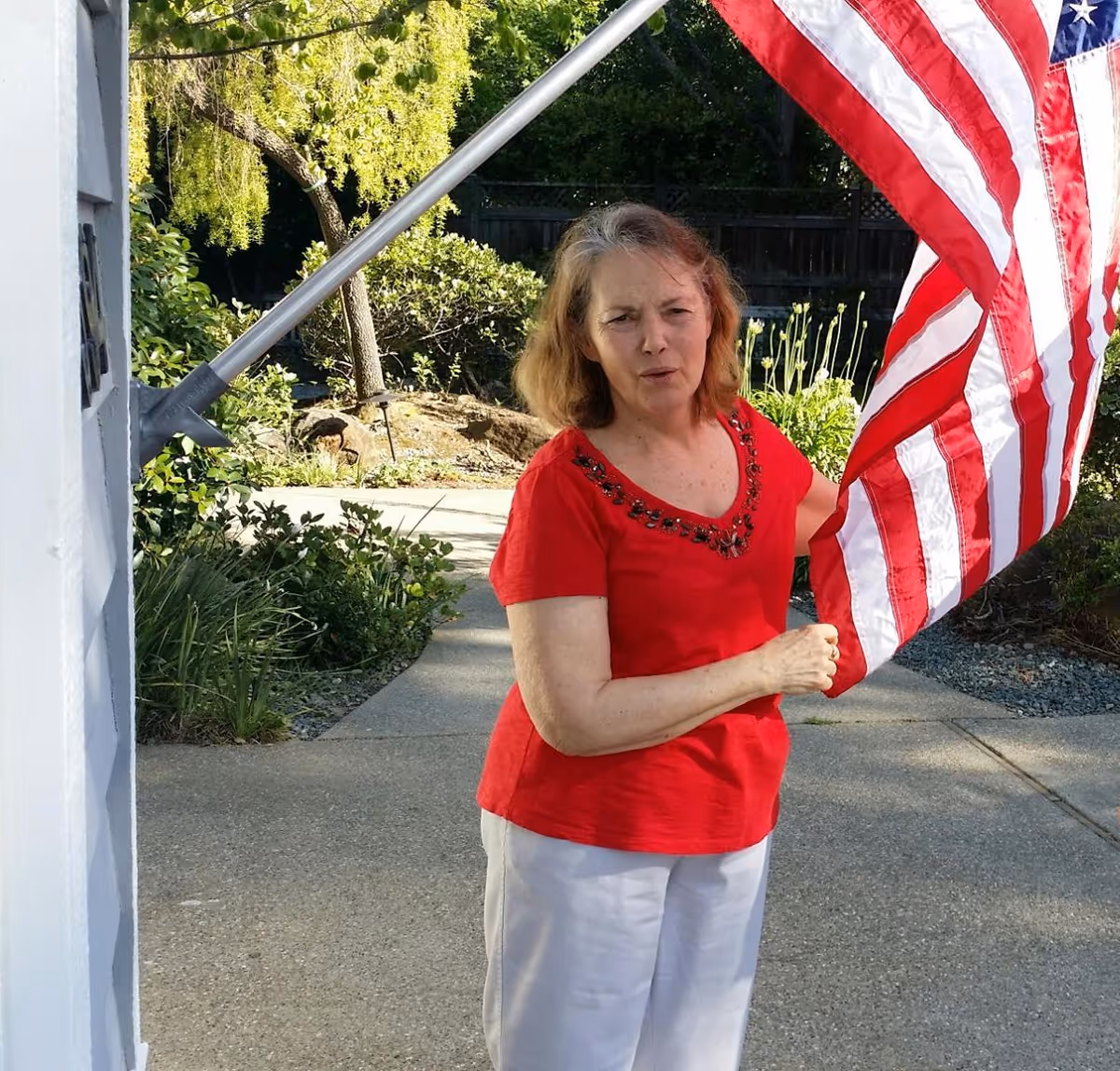 A woman wearing a red shirt and white pants is standing outside near a house, holding an American flag mounted on a pole. There are green bushes, trees, and a concrete pathway in the background.