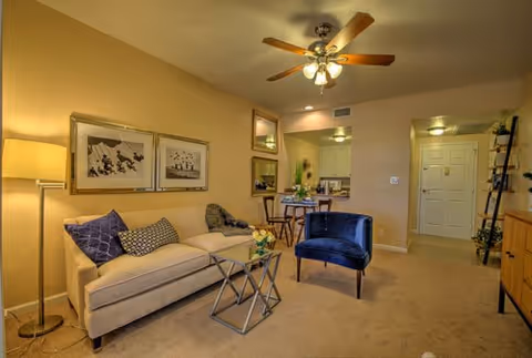 A cozy living room with a beige sofa adorned with patterned cushions, a blue upholstered chair, a glass-top coffee table with flowers, a floor lamp, and framed artwork on the wall. The room has beige walls and carpet, a ceiling fan with lights, and an open view into a small dining area and kitchen. The entrance door is visible in the background.