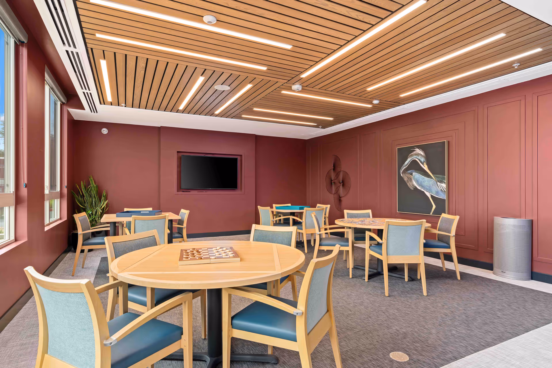 A well-lit common room with multiple round wooden tables and chairs arranged for group activities. The walls are painted a deep red, adorned with a framed painting of a bird and a decorative wall sculpture. The ceiling features wooden slats with integrated linear lighting. Large windows on one side allow natural light to enter the room.