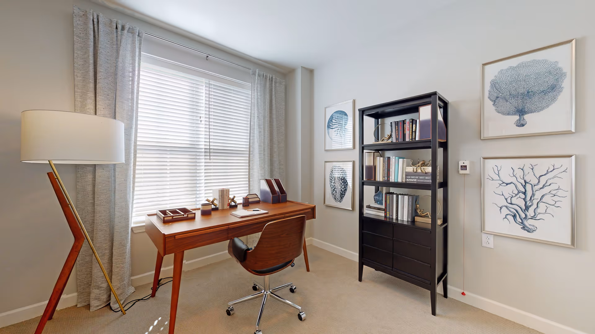 A bright and modern office space with a wooden desk and a swivel chair on wheels. The desk is positioned in front of a window with white blinds and light gray curtains. To the left of the desk is a tall floor lamp with a white lampshade and a wooden and brass stand. On the right side of the room, there is a black bookshelf filled with books and decorative items. The walls are decorated with framed blue and white coral artwork.