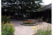 A peaceful outdoor courtyard with a circular flower bed in the center surrounded by a brick-paved area. There are various yellow flowers and greenery around the edges, with a bench visible near the building walls in the background.