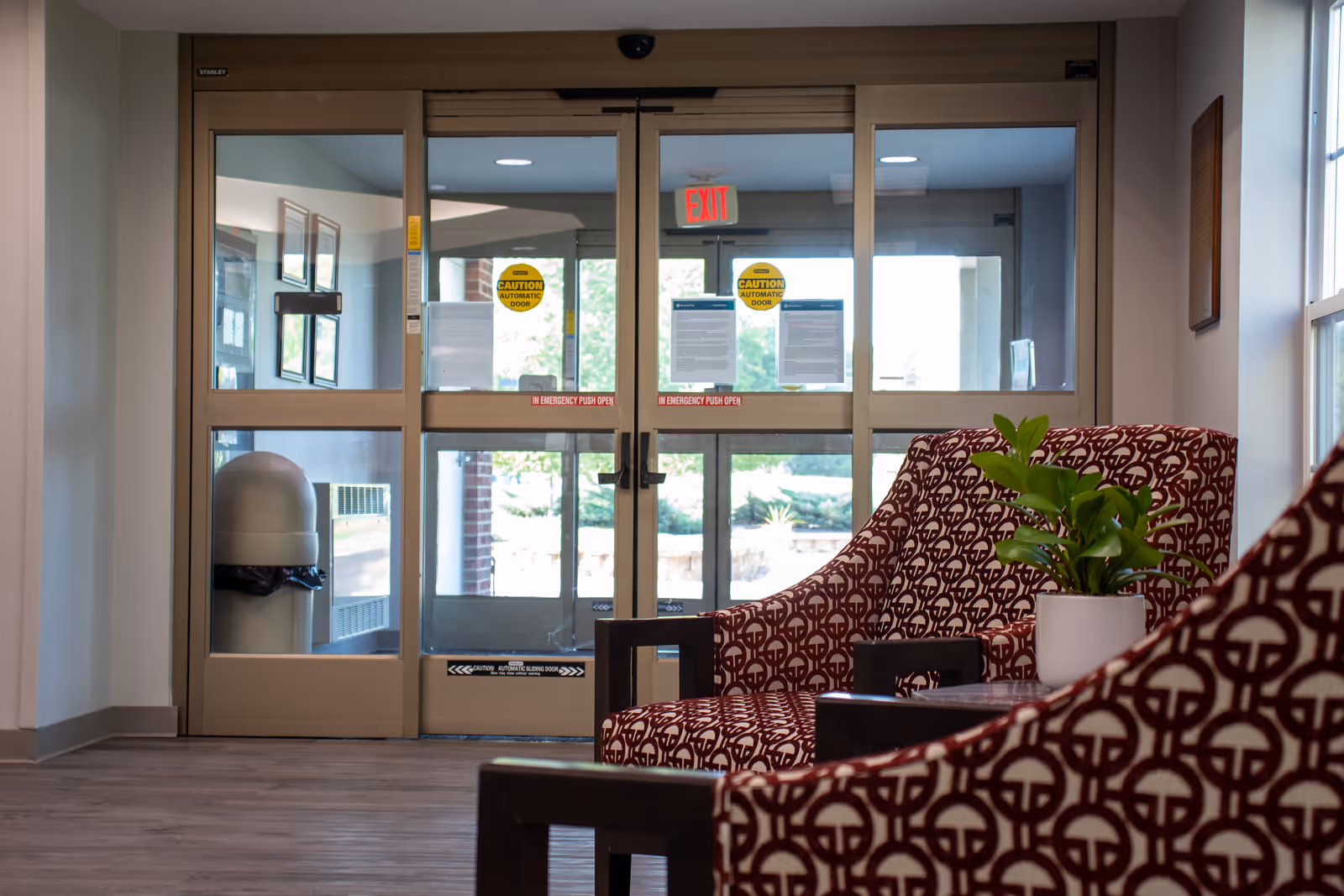 View of a seating area inside a building near automatic glass double doors. Two patterned armchairs with a small table and a potted plant are visible in the foreground. Outside the doors, some greenery and part of the building exterior can be seen.