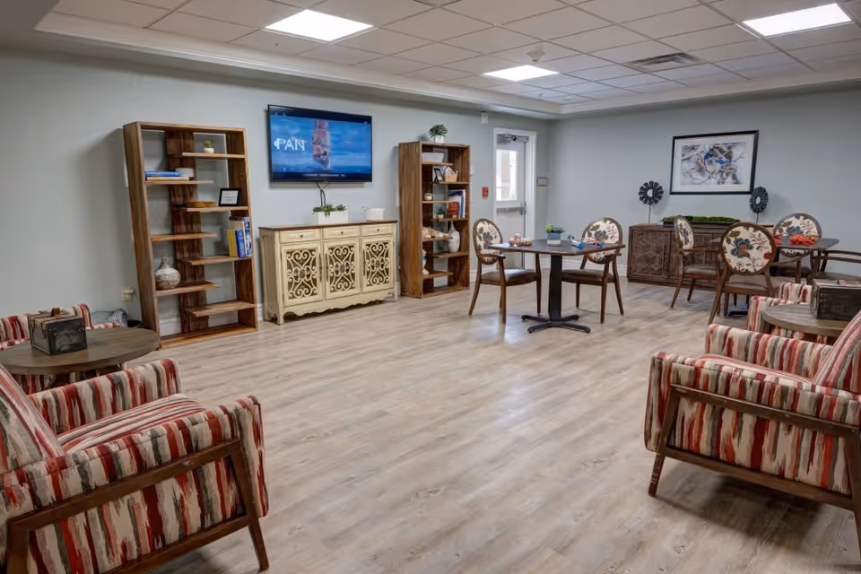 A spacious common area with light wood flooring and light blue walls. The room features two striped armchairs with wooden frames, two round tables with floral upholstered chairs, wooden bookshelves, a decorative cabinet, a wall-mounted TV, and a framed artwork. The ceiling has recessed lighting, and there is a door leading outside or to another room.