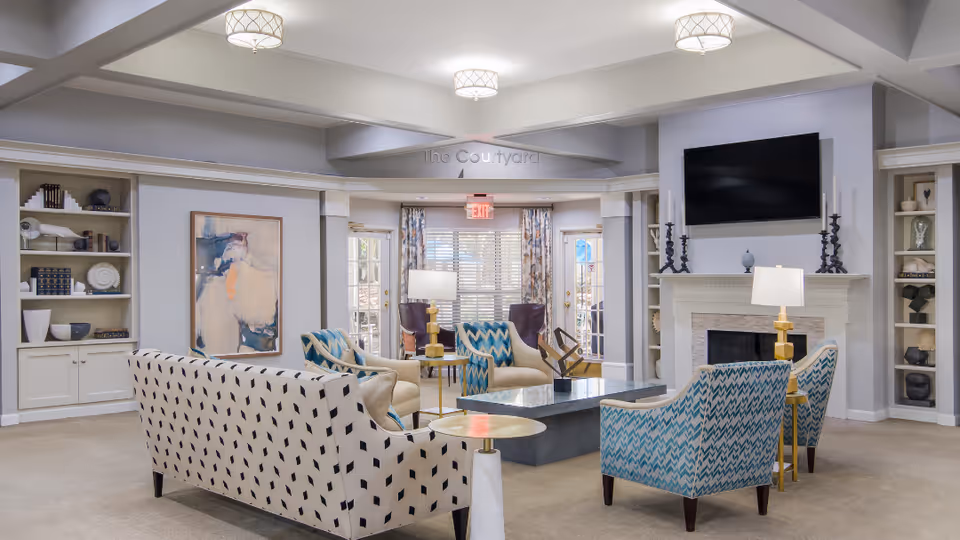 A spacious and well-lit living room area with patterned sofas and armchairs arranged around a rectangular coffee table. The room features built-in shelves with decorative items, a fireplace with a mounted flat-screen TV above it, and large windows with blinds letting in natural light. There are also two lamps on side tables and a painting on the wall.