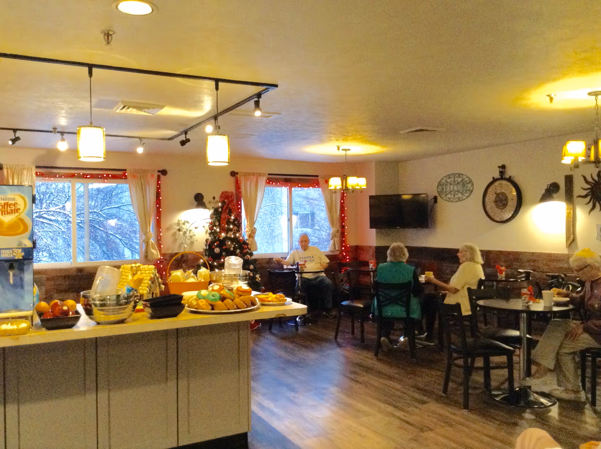 A cozy dining area in a retirement community decorated for the holidays with a Christmas tree and red lights. Several elderly people are seated at tables enjoying snacks and drinks. A counter in the foreground displays various food items including donuts, fruit, and a Coffee-mate dispenser.