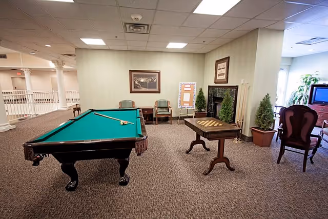 A recreational room with a pool table and a chess/checkers table. The room has carpeted flooring, light-colored walls, framed pictures, and several chairs. There are potted plants near a fireplace and a television in the background.
