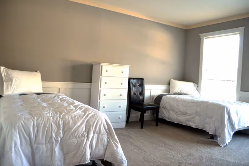 A bedroom with two single beds, each with white bedding and pillows. Between the beds is a white chest of drawers and a black cushioned chair. There is a window on the right side letting in natural light, and the walls are painted gray with white trim.