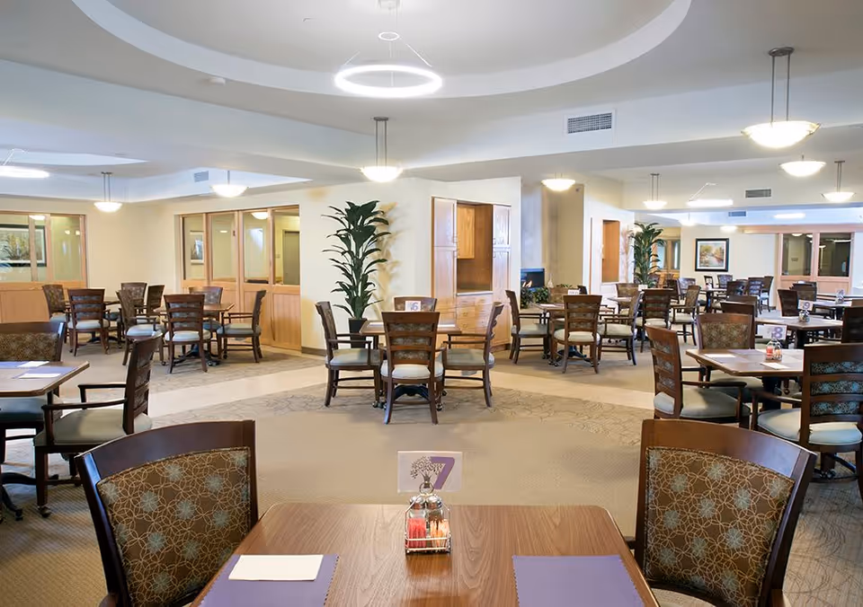 A spacious dining room with multiple wooden tables and chairs arranged neatly. Each table has placemats, napkins, and a small condiment holder. The room is well-lit with modern ceiling lights and decorated with potted plants and framed artwork on the walls.