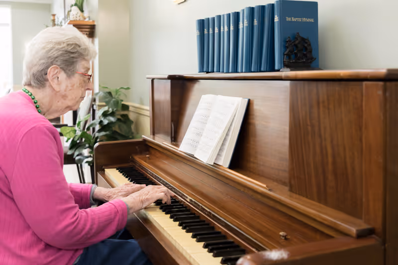 An elderly woman wearing a pink sweater and green necklace is playing a wooden piano with sheet music open. There are blue books and a small ship model on top of the piano, and a plant and furniture are visible in the background.