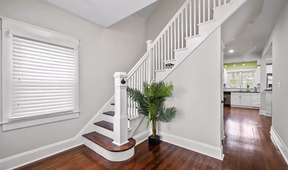Interior view of a home showing a wooden staircase with white railings, a large window with white blinds, a potted green plant at the base of the stairs, and a hallway leading to a kitchen with white cabinets and a green accent wall.