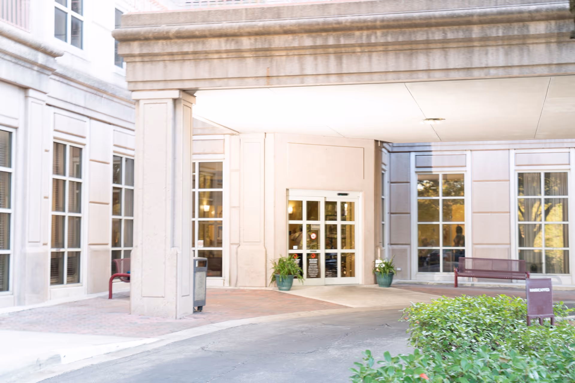 Entrance area of a healthcare facility with automatic sliding glass doors, potted plants on either side of the entrance, benches, and large windows on the building exterior.
