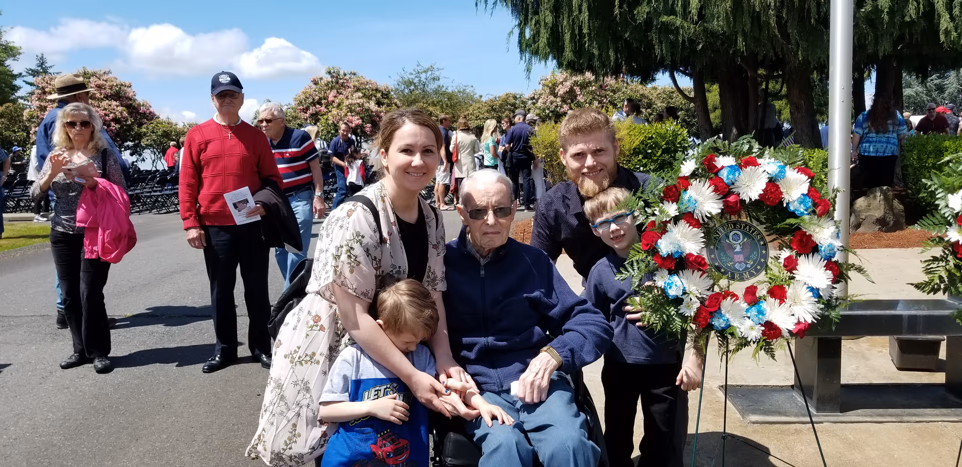 A group of people outdoors on a sunny day, including an elderly man in a wheelchair surrounded by family members, with a patriotic wreath featuring red, white, and blue flowers nearby. Other people are walking and standing in the background along a paved path with trees and bushes.