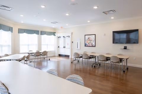 A bright, spacious room with multiple tables and chairs arranged for group seating. Large windows with blue valances allow natural light to fill the room. A flat-screen TV is mounted on the wall, and a colorful framed portrait hangs nearby. The room has wooden flooring and white walls, creating a clean and welcoming atmosphere.